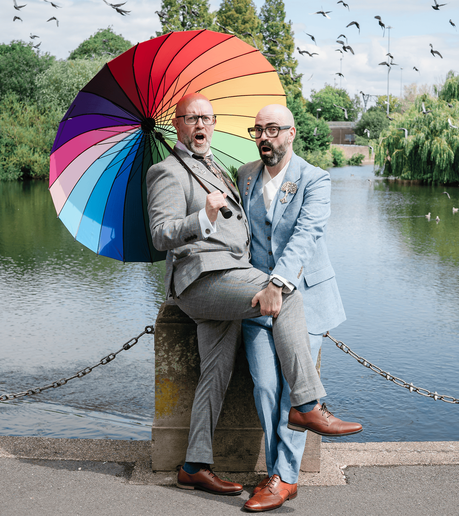 Vivid rainbow umbrella held by two men in suits near a river, with birds flying overhead and lush gr.