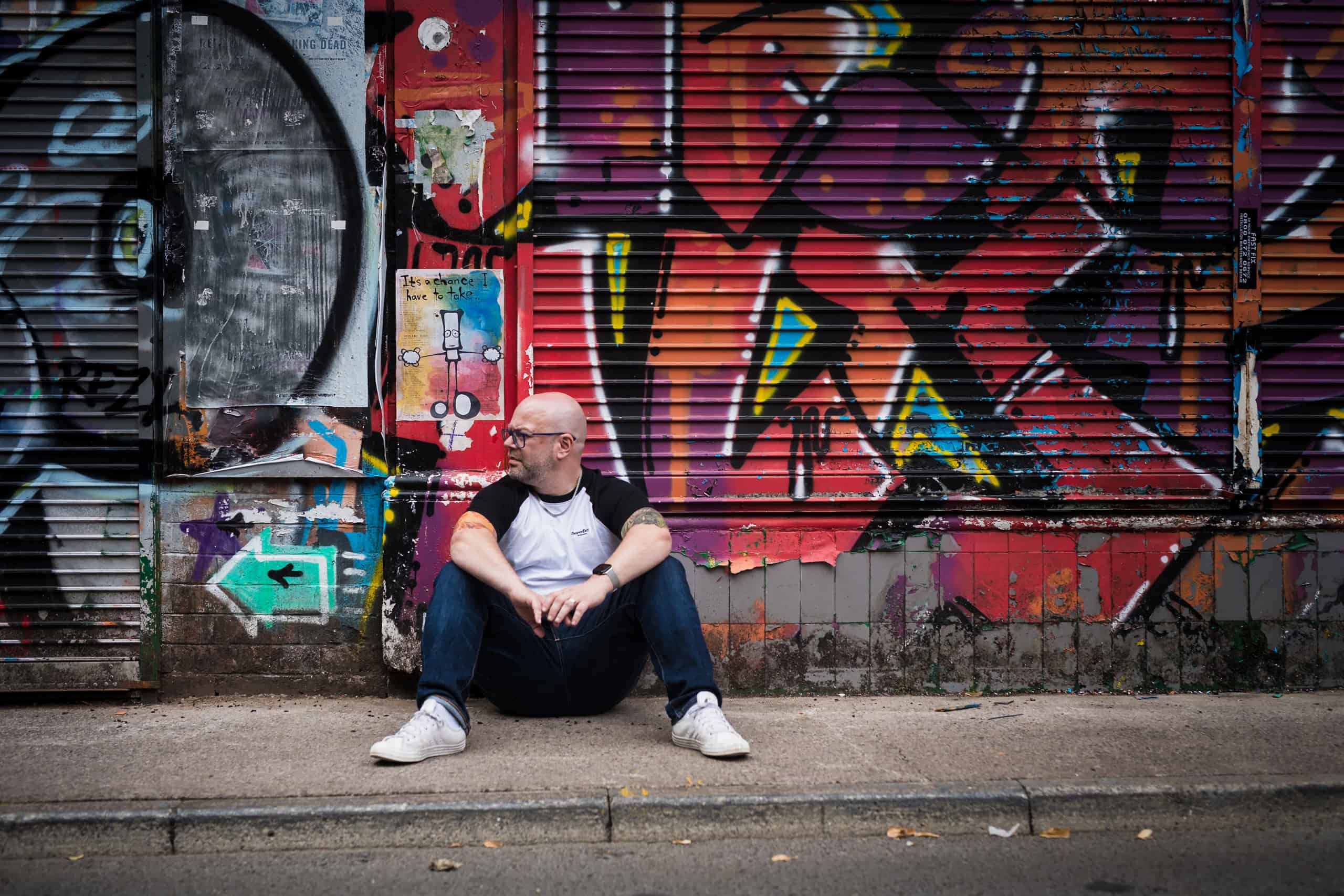 Urban graffiti wall with man sitting on sidewalk, colourful street art background.