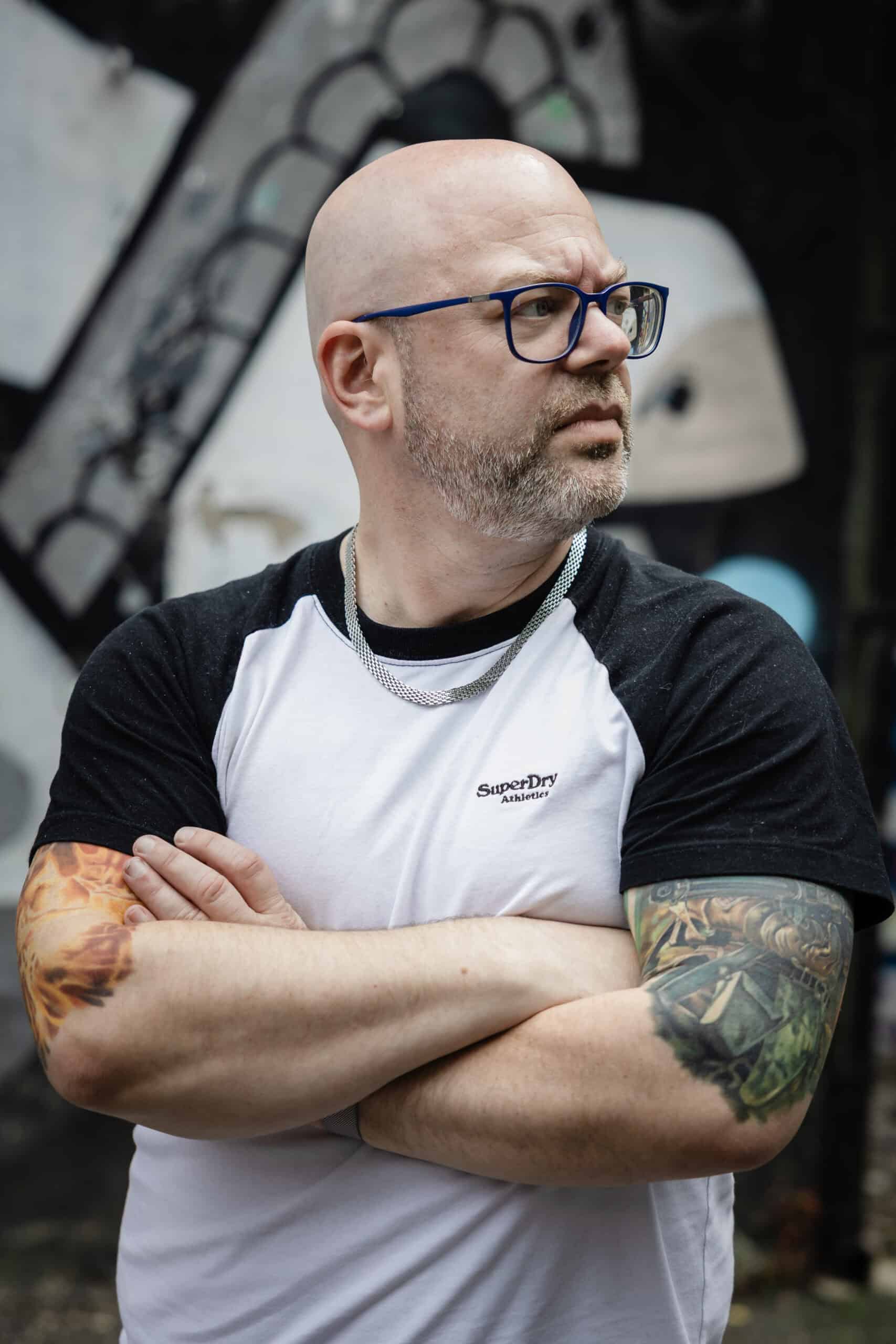 Portrait of Paul Phipps-Williams with tattoos and glasses in urban graffiti background.