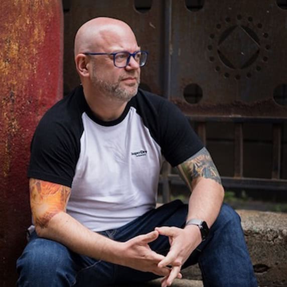 Portrait of Paul Phipps-Williams, photographer with tattoos and glasses, sitting outdoors.