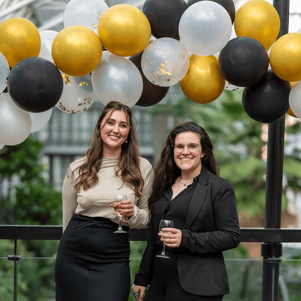 Two women smiling with champagne glasses at a celebration with gold, black, and white balloons.