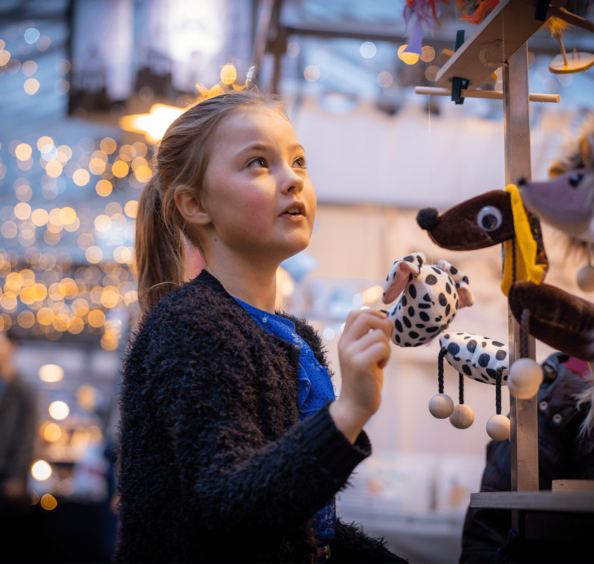 Child browsing plush animal toys at a Christmas market stall, with festive lights in the background.