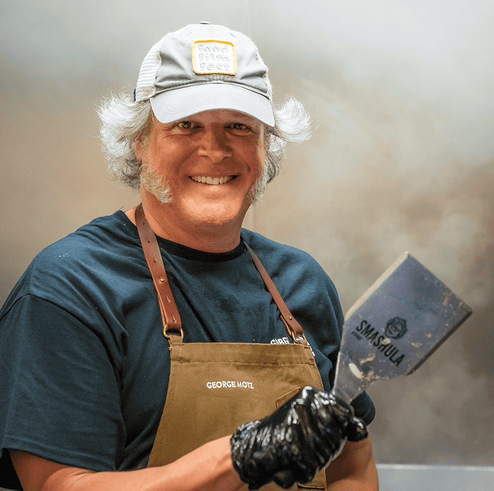 Paul Phipps-Williams smiling with a spatula in hand, wearing a cap and apron, capturing culinary pho.