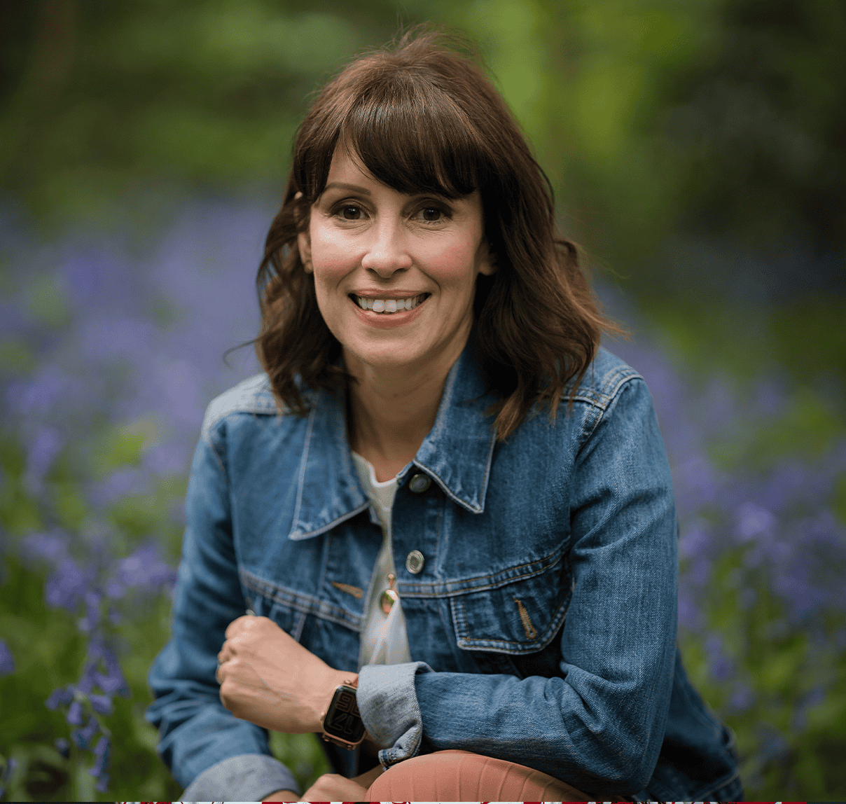 Portrait of a woman in a denim jacket outdoors with purple flowers in the background.