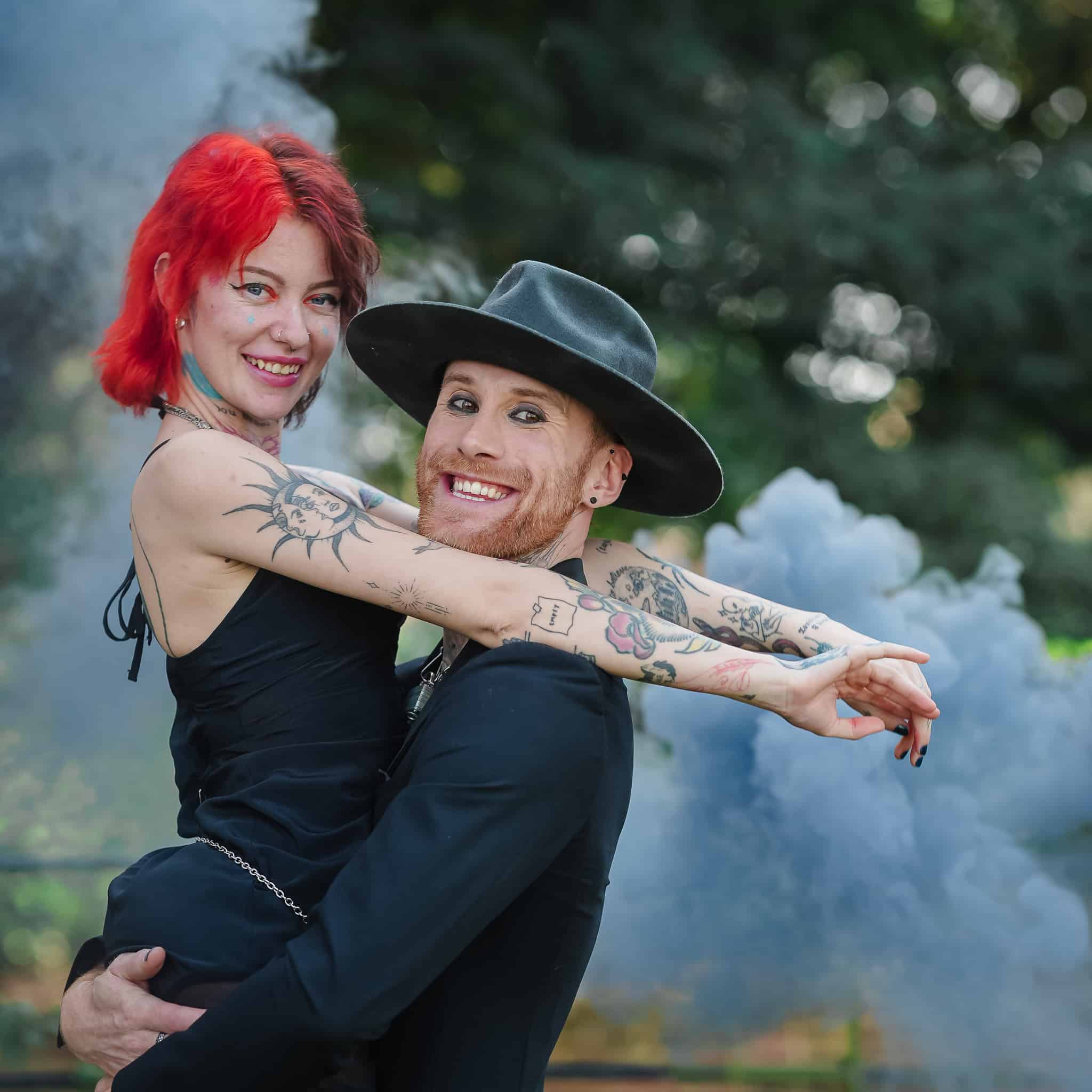 Red-haired woman and tattooed man embracing outdoors with smoke in background.