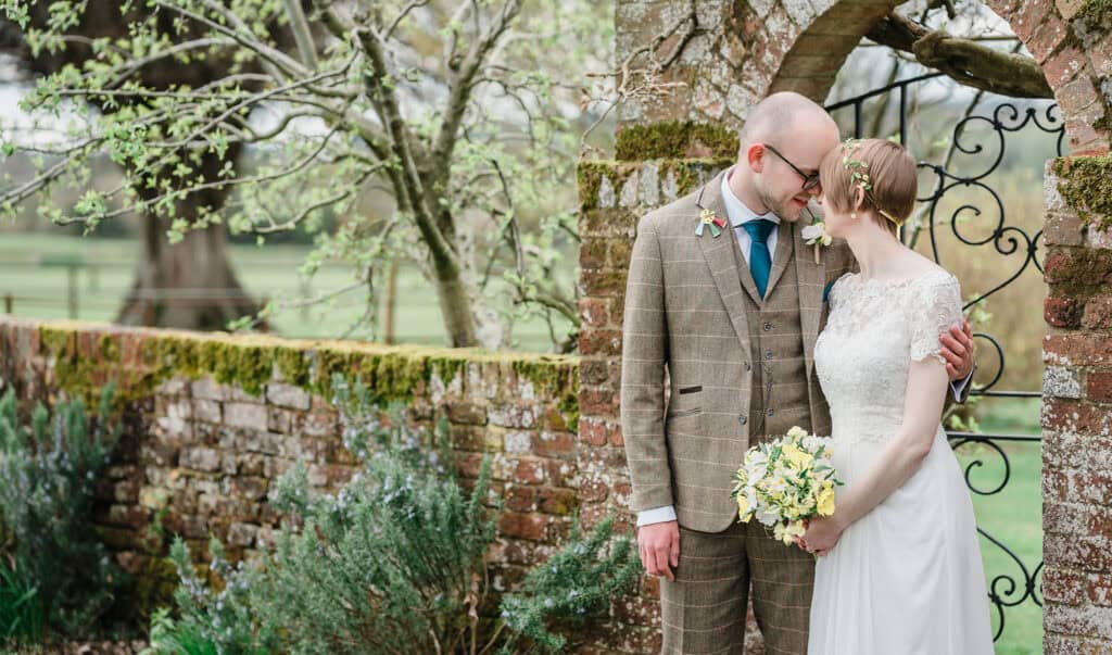 Bride and groom sharing a tender moment in a lush garden with historic brick archway.