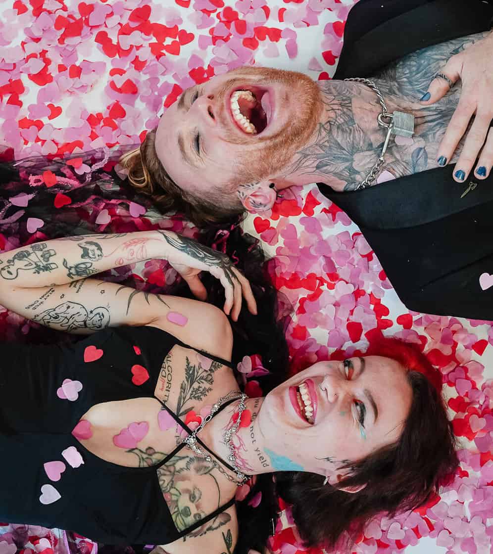 Happy couple with tattoos lying on heart-shaped confetti floor, smiling and enjoying a joyful moment.