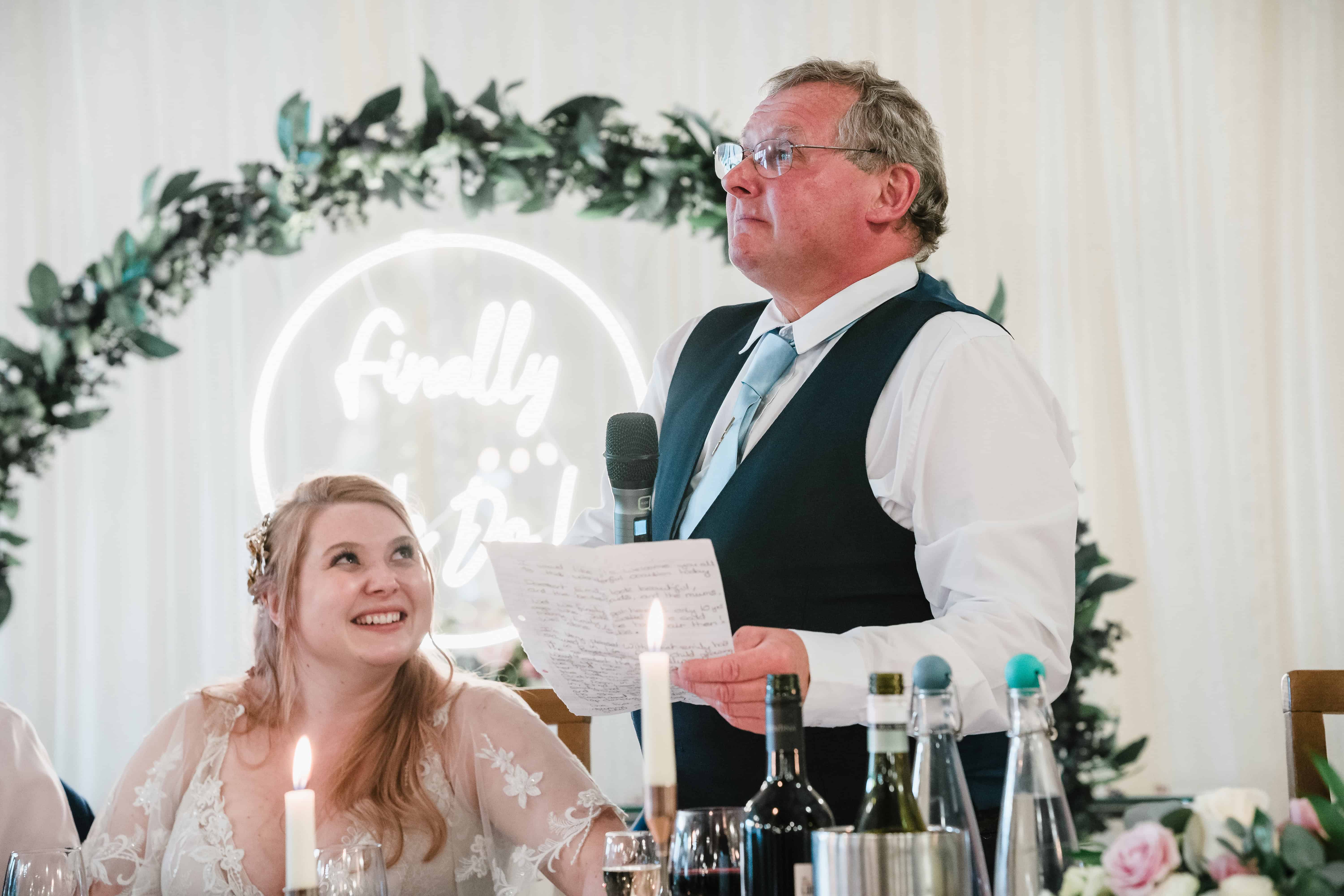 a bride looks at her father with pride as he gives his speech
